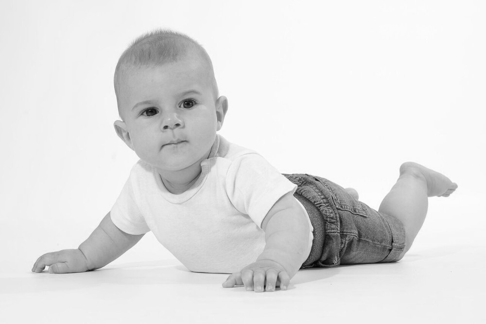 Ein Baby liegt auf dem Bauch und schaut in die Kamera, gekleidet in ein weißes T-Shirt und kurze Jeans.