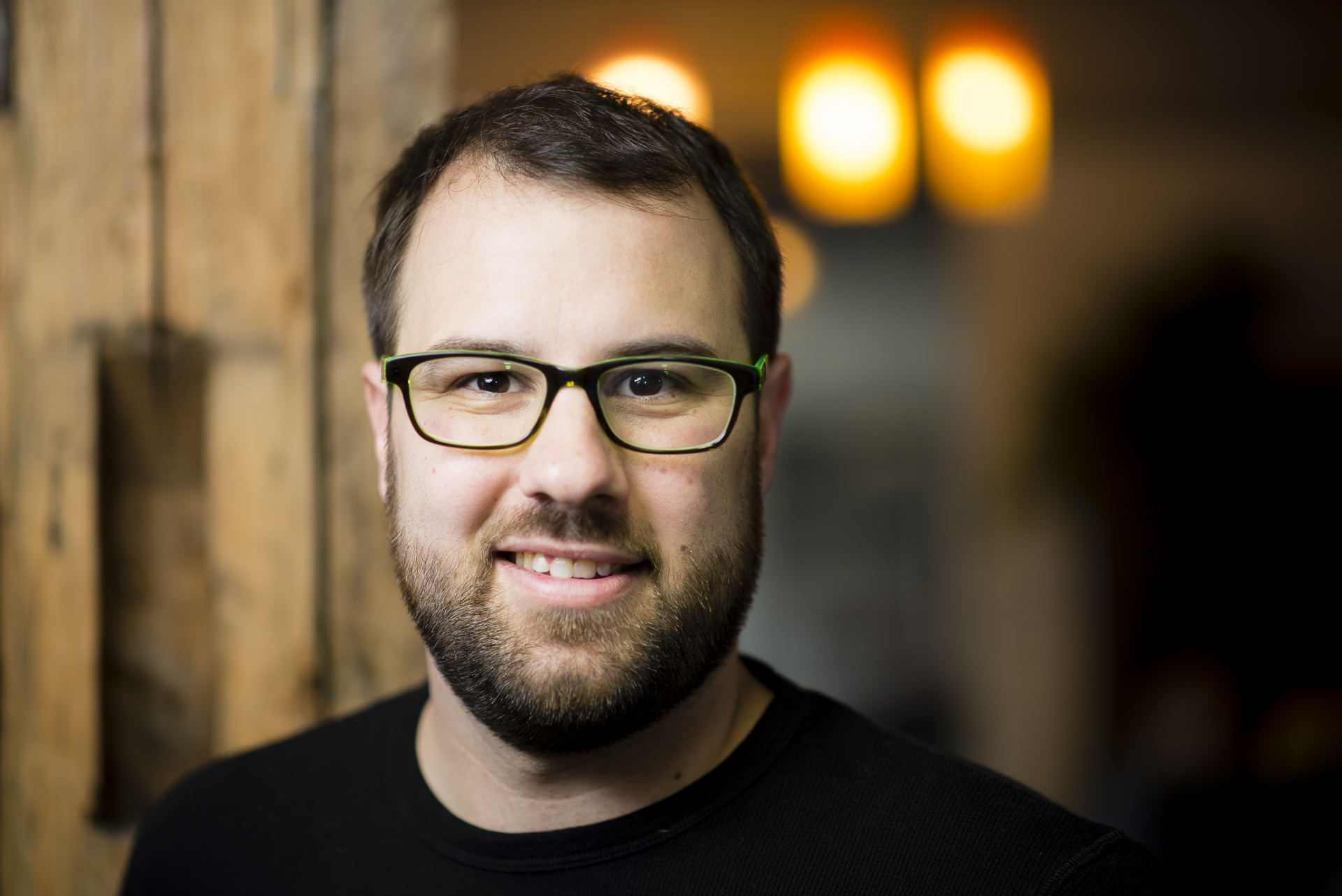 Ein Mann mit Brille und Bart lächelt vor einem unscharfen Hintergrund mit warmen Lichtern.